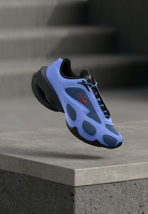 Niebiesko-czarny sportowy but z czerwonym logo Nike, czarnymi sznurowadłami i teksturowanymi panelami, unoszący się nad betonowym krawężnikiem, w tle schody.