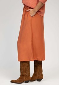 Jupe mi-longue de couleur rouille avec une texture lisse, dotée d'une taille élastique et de poches sur les côtés, portée avec des bottes de cowboy en daim marron.