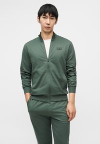 Πράσινο μπουφάν με φερμουάρ και όρθιο γιακά, κατασκευασμένο από λείο ύφασμα, με μικρό μαύρο λογότυπο στο αρι στερό μέρος του στήθους, συνδυασμένο με παντελόνι στον ίδιο χρωματικό τόνο.