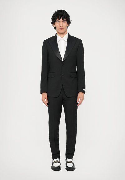 Schwarzer Anzug mit satinierte Revers, kombiniert mit einem weißen Hemd. Die Hose passt zum Blazer. Schwarze Schuhe mit goldenen Akzenten.