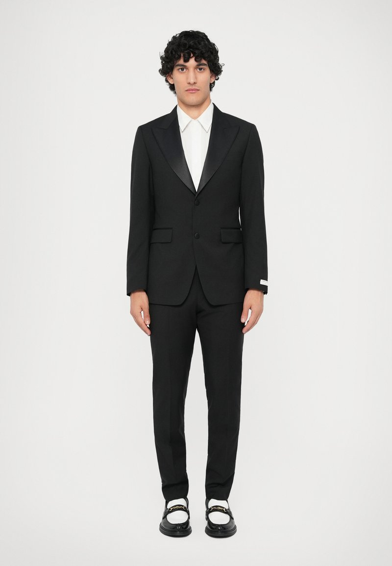 Čierny oblek so saténovým špicatým klopom, v kombinácii s bielou košeľou. Nohavice zodpovedajú saku. Čierne topánky so zlatými akcentmi.