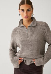 Lys grå strikket sweater med kvart-zip krave, ribbet tekstur og lange ærmer. Har sidespalter ved kanten.