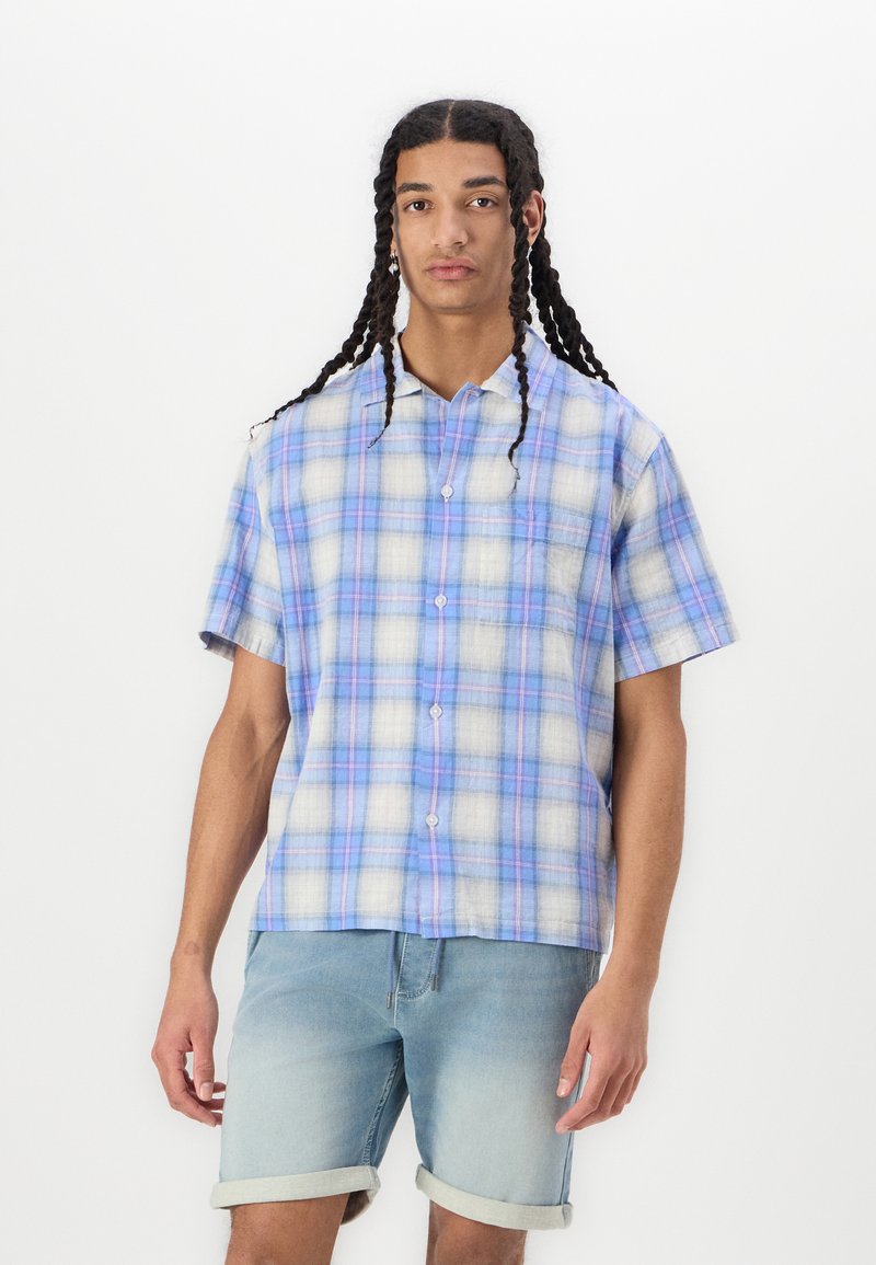 Short-sleeve plaid shirt in light blue, white, and purple with a button-down front and pocket. Worn with light denim shorts.