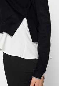 Czarny sweter z dzianiny z falbaną u dołu, noszony na białej bluzce; ozdobiony srebrną nicią przy zakończeniach rękawów i dolnym brzegu.