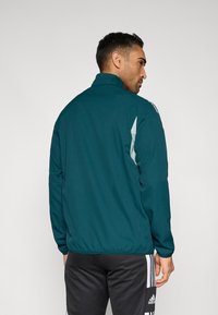 Sinivihreä tuulitakki, jossa korkea kaulus, joustavat hihansuut ja kapea istuvuus. Takissa on vaaleanharmaat sivukoristeet ja sileä, kevyt kangas.