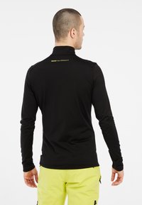Zwarte langesleeve top met een hoge kraag en een gladde textuur. Heeft een klein geel logo op de achterkant, gecombineerd met felgele broek.