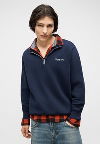 Νεαρός άνδρας που φοράει μπλε σκούρο φούτερ με φερμουάρ μέχρι τη μέση πάνω από κόκκινο και μαύρο πουκάμισο καρό, με μπλε τζιν, στέκεται μπροστά σε απλό φόντο.