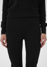 Pantaloni neri aderenti realizzati in tessuto liscio, con vita alta. Indossati con un maglione a maglia scura e strutturata. Mani visibili ai lati.