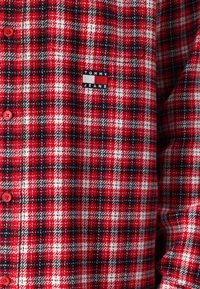 Raudona ir juoda languota flanelinė marškiniai su baltais akcentais, su blizgančiomis raudonomis sagomis ir mažai "TOMMY JEANS" etikete ant krūtinės.