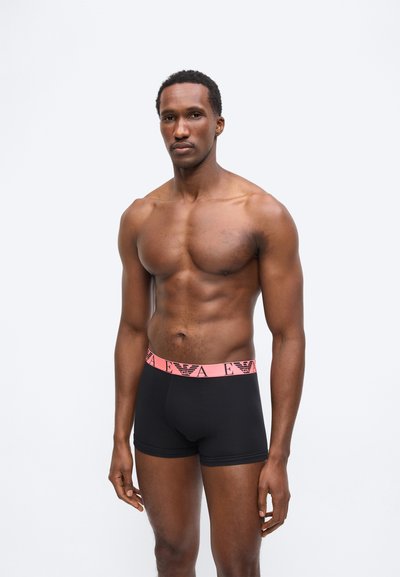 Ευθυτενής άντρας όρθιος, φορώντας μαύρα μποξεράκια με ροζ ζώνη μέσης που φέρει γραμμένο το εμπορικό σήμα και λογότυπο, σε απλό φωτεινό φόντο.