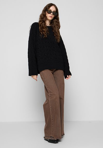 Femme portant un pull noir en grosse maille oversize, un pantalon large marron, des chaussures noires et des lunettes de soleil foncées, debout devant un fond clair.