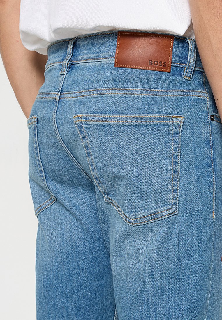 Jean en denim bleu clair avec une patch en cuir brun portant le logo BOSS sur la taille. Les caractéristiques incluent des poches arrière et des détails de couture visibles.
