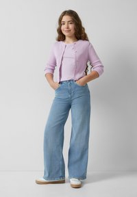 Helllila gerippte Strickjacke mit Knöpfen, getragen über einem pastellfarbenen Oberteil, kombiniert mit weit geschnittenen hellblauen Jeans und weißen Turnschuhen mit einer sandfarbenen Sohle.
