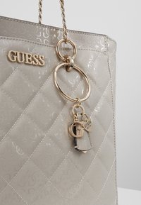 Sac à main matelassé beige brillant Guess avec poignée en chaîne dorée, logo Guess doré et porte-clés dorés comprenant un cœur et la lettre G.