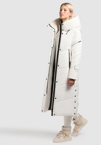 Ilgas baltas pūkinis paltas su aukštu apykakle, su juodu užtrauktuku ir spaustukais. Prisegtas prigludęs vidinis sluoksnis. Dėvima su smėlio spalvos auliniais batais.