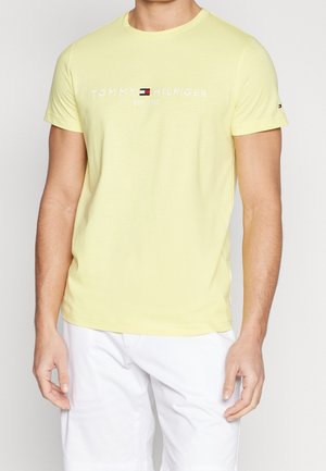 Mann trägt ein hellgelbes Tommy Hilfiger T-Shirt mit Logo und weiße Shorts, steht mit entspannten Armen an den Seiten.