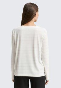 Haut blanc à manches longues avec des fines rayures horizontales ; tissu doux, encolure ronde et fentes sur les côtés à l'ourlet.