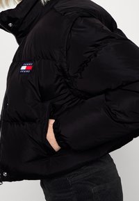 Sort polstret jakke med lynlås, med tilbagetrukne ærmer, sidelommer og et farverigt Tommy Jeans-logo på venstre side.