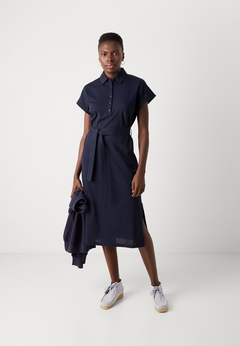 Granatowa sukienka do kolan z kołnierzykiem, krótkimi rękawami, związaną w talii paskiem i bocznymi rozcięciami. Para jasnych butów.