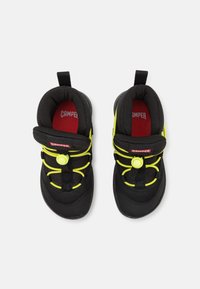 Zapatos infantiles negros con parte superior de tela texturizada, que presentan cordones elásticos amarillos brillantes y una plantilla roja con el logo de "Camper".