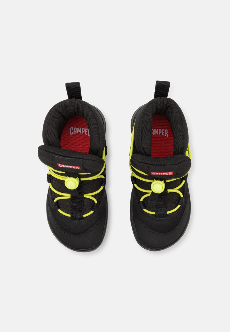 Scarpe nere per bambini con tomaia in tessuto texturizzato, dotate di vivaci lacci elastici gialli e una soletta rossa con il logo "Camper".