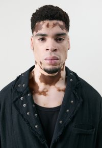Schwarze Jacke mit ausgefransten Kanten und silbernen Nieten, über einem eng anliegenden schwarzen Oberteil getragen, von einer Person mit auffälligen Hautmustern.