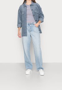 Camisa de pana azul oversize superpuesta sobre una camiseta a rayas azul claro y rosa, combinada con vaqueros de pierna recta de lavado claro y zapatillas blancas.
