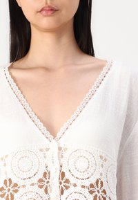 Blouse blanche à col en V avec une bordure en dentelle. Présente un motif au crochet avec des motifs circulaires et une fermeture à boutons sur le devant. Tissu texturé.