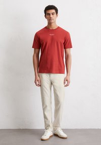 Red cotton t-shirt with short sleeves and crew neckline, featuring a small logo in white at the center. Worn with light beige pants and white sneakers.