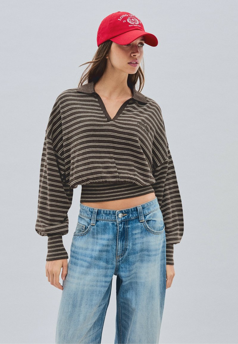 Femme portant une casquette rouge, un crop-top à manches longues ample rayé marron et blanc, et un jean taille haute bleu clair, debout devant un fond neutre.