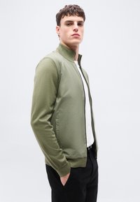 Νέος άνδρας που στέκεται με τα χέρια στις τσέπες, φορώντας πράσινο μπουφάν με φερμουάρ πάνω από λευκό πουκάμισο και μαύρο παντελόνι, κοιτάζοντας ελαφρώς προς το πλάι.