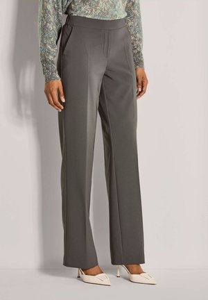 Vrouw in een grijze broek met wijde pijpen, een abstract gestreepte lange mouwtop en witte puntige hakken, staande tegen een neutrale achtergrond.