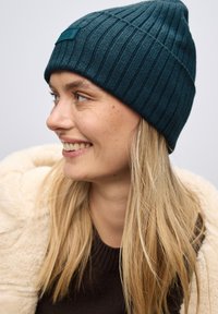 Tyrkysový pletený beanie s texturovaným povrchem a obdélníkovou nášivkou na přední straně, který má na sobě osoba s dlouhými blond vlasy.