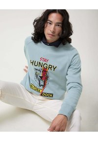 Sudadera de color azul claro con un gráfico de un tigre y el texto "STAY HUNGRY" en rojo y amarillo. Combinada con pantalones blancos; presenta puños acanalados.