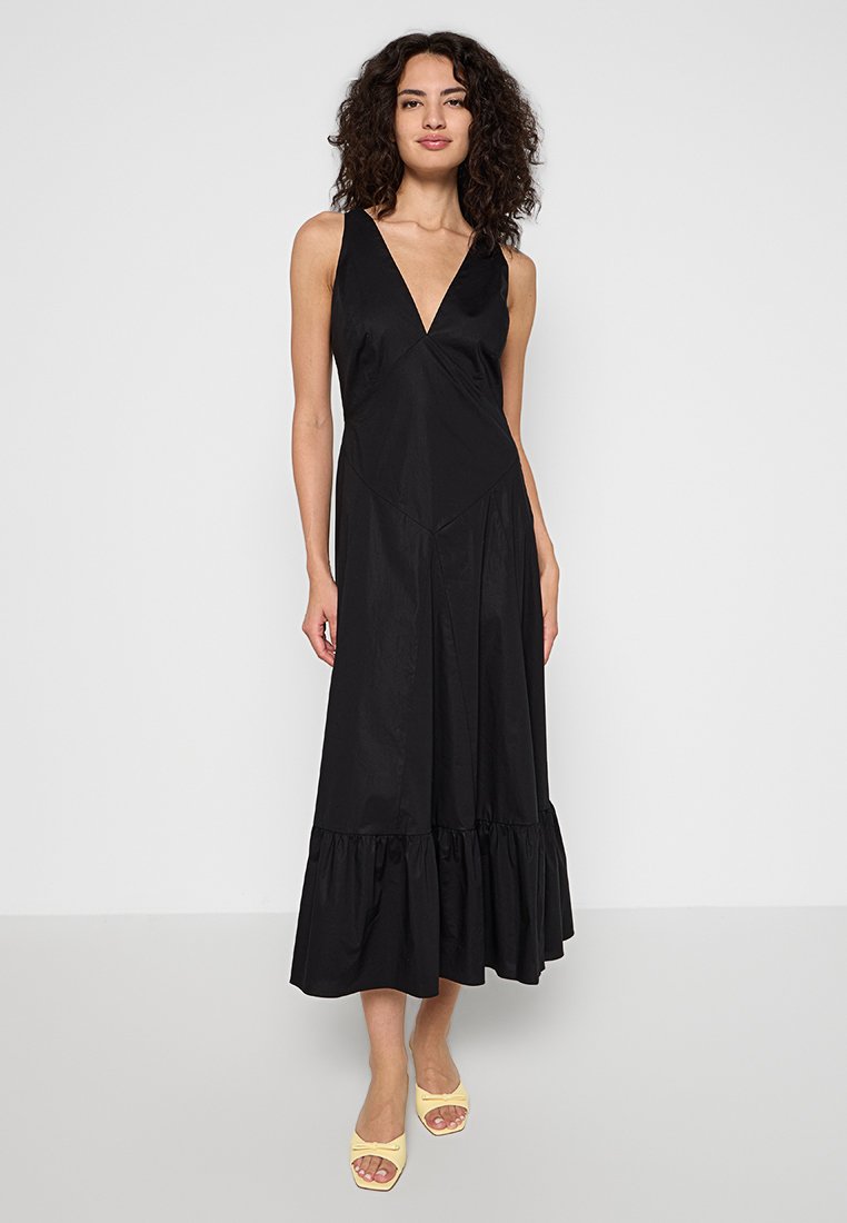 Czarna sukienka bez rękawów z głębokim dekoltem w kształcie litery V, rozkloszowanym dołem i teksturowanym wykończeniem. Modelka nosi żółte sandały. Prosta, elegancka sylwetka.