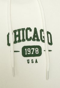 Sudadera crema con texto bordado en verde: "CHICAGO 1978 USA." Presenta cordones con puntas de plástico y una textura de tela suave.