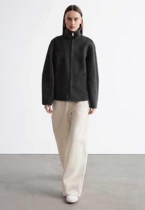 Vrouw die naar voren loopt en een donkergrijze jas met rits draagt, roomkleurige wijde pijpen broek, witte schoenen en gouden creolen tegen een effen achtergrond.