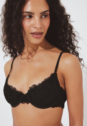 Vrouw met krullend donker haar, gekleed in een zwarte kanten BH, kijkt iets opzij tegen een effen witte achtergrond.