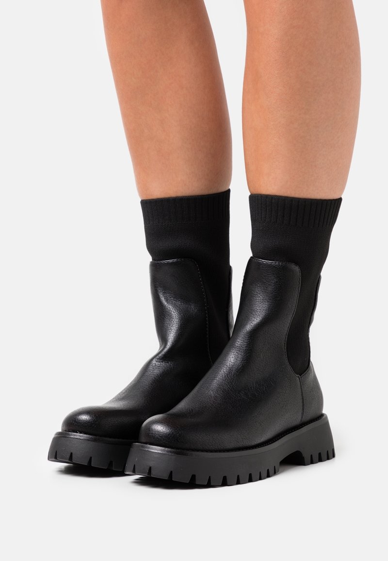 Jambes portant des bottines en cuir noir avec des semelles épaisses et des parties supérieures côtelées noires, de type chaussette, sur un fond uni.