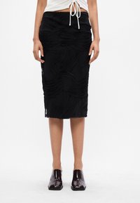 Femeie îmbrăcată cu o fustă texturată neagră până la genunchi, un top alb scurt cu șnur și pantofi slip-on lucioși de culoare maro închis, stând pe un fundal alb.