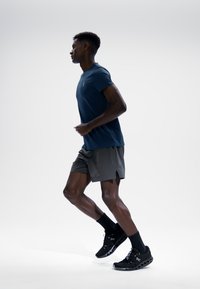 Athlet in einem dunkelblauen Kurzarmhemd und grauen Laufshorts, der schwarze Sportschuhe und schwarze Socken trägt, läuft vor einem einfachen Hintergrund.