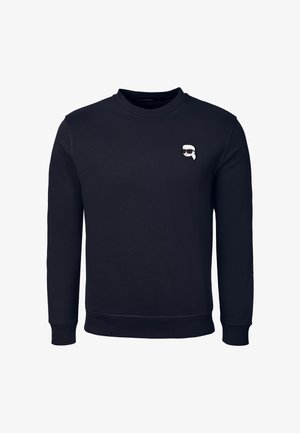 Zwarte sweatshirt met lange mouwen en ronde hals, met een klein witte en zwarte cartoon gezicht met een zonnebril op de linkerborst.