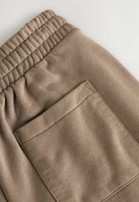 Beige sweatpants har en ribbet elastisk talje og en lomme. Stoffet har en blød tekstur og en glat overflade.