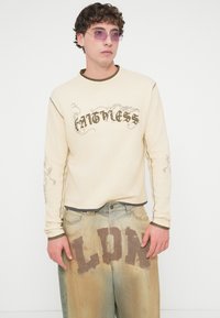 Μπεζ μακρυμάνικο πουκάμισο με καφέ κείμενο και διακοσμητικές λεπτομέρειες στα μανίκια, συνδυασμένο με ξεθωριασμένο μπλε τζιν παντελόνι με έντονη επιγραφή "LDN".