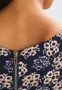 Marinblommig klänning med rund halsringning som har beige, röd och grön mönster, samt en synlig metalldragkedja på baksidan.