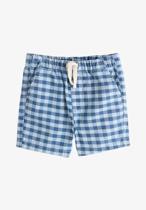 Młody chłopiec uśmiecha się, stoi z rękami w kieszeniach, nosi granatowy T-shirt, niebieskie szorty w kratkę, ciemne skarpetki i białe trampki.