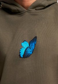 Olíva zöld kapucnis felső, amelyen egy kék pillangó grafika látható, részletes szárnyakkal, a mellkas bal oldalán elhelyezve.
