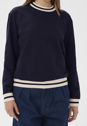 Frau trägt einen marineblauen Pullover mit weißen Querstreifen am Rundhalsausschnitt, an den Bündchen und am Saum, kombiniert mit hoch taillierten dunkelblauen Jeans, vor einem neutralen Hintergrund.