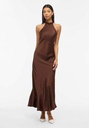 Kvinne iført en ermeløs brun satengkjole med halterneck og beige spisse hæler, stående mot en enkel hvit bakgrunn.