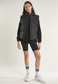 Giovane donna che indossa un gilet imbottito nero, una maglietta a maniche lunghe nera, pantaloncini da ciclismo neri, calzini bianchi con strisce nere e scarpe da ginnastica bianche in piedi.